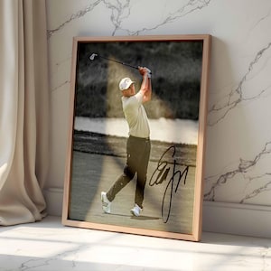 May include: A framed wall art print featuring a golfer in a light-colored shirt, dark pants, and a white cap mid-swing on a golf course. The image includes a prominent black signature overlaying the lower right portion of the photograph.