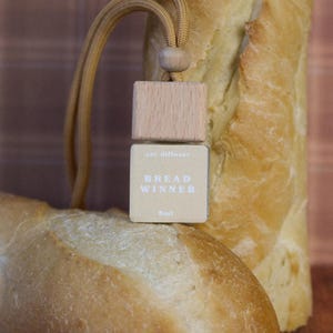 Puede incluir: Un ambientador para coche con tapa de madera y etiqueta beige con "BREAD WINNER". El ambientador está suspendido por un cordón marrón y se coloca delante de dos barras de pan. La etiqueta también indica 8ml.