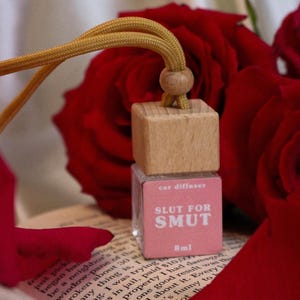 Puede incluir: Un ambientador para coche con una parte superior de madera y una etiqueta rosa que dice "SLUT FOR SMUT". El ambientador cuelga con un cordón dorado y está rodeado de pétalos de rosa rojos y rosas.