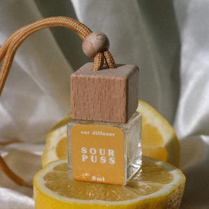 Pode incluir: Um pequeno frasco difusor de carro de vidro quadrado com tampa de madeira clara e cordão bege para pendurar. O rótulo é amarelo com texto branco que diz car diffuser, SOUR PUSS e 8ml. O frasco está posicionado sobre um limão amarelo cortado.