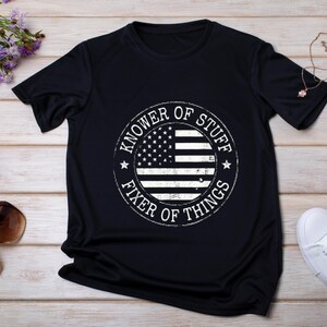 Peut inclure: Un t-shirt bleu marine avec un motif blanc représentant un drapeau américain et le texte "Knower of Stuff, Fixer of Things" dans un motif circulaire.