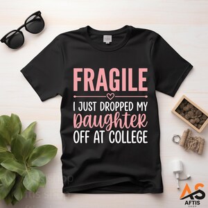 Peut inclure: T-shirt noir avec l'inscription "FRAGILE I JUST DROPPED MY daughter OFF AT COLLEGE" en rose et blanc. Le t-shirt a un col rond et des manches courtes. Des lunettes de soleil noires et une petite plante sont visibles.