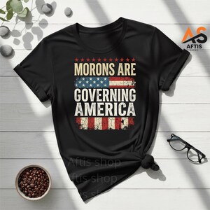 Peut inclure: T-shirt noir avec l'inscription blanche "MORONS ARE GOVERNING AMERICA" et un drapeau américain vieilli. Le t-shirt a un col rond et des manches courtes. Le texte est en gras, sans empattement.