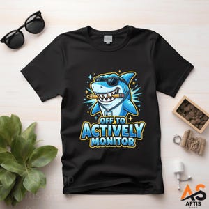 Peut inclure: T-shirt noir avec un requin de dessin animé portant des lunettes de soleil et tenant un crayon dans sa bouche. Le texte "OFF TO ACTIVELY MONITOR" est en bleu et jaune. Des lunettes de soleil et une petite plante sont également visibles.