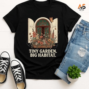 Peut inclure: Un t-shirt noir à manches courtes orné d'une illustration représentant une fenêtre arquée avec des volets ouverts et une jardinière remplie de fleurs colorées et de papillons. Sous l'image, le texte indique : TINY GARDEN. BIG HABITAT.