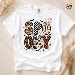 Peut inclure: Un t-shirt blanc à manches courtes avec le mot SPOOKY écrit en grandes lettres à motifs. Les lettres présentent des imprimés léopard, des damiers noirs et blancs, des citrouilles orange et des fantômes. Des toiles d'araignées, des araignées et des chauves-souris noires entourent le texte.