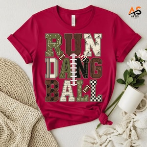 Peut inclure: Un t-shirt rouge à manches courtes avec le texte RUN DANG BALL en grandes lettres majuscules. Les lettres sont remplies de motifs sur le thème du football, incluant des schémas de jeu, des casques et des carreaux, avec un lacet de ballon.