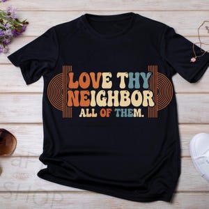 Peut inclure: Un t-shirt noir avec l'inscription "LOVE THY NEIGHBOR ALL OF THEM" en orange, beige et bleu. Le texte est encadré de lignes courbes orange et marron. Le t-shirt est présenté sur une surface en bois clair.
