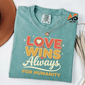 Peut inclure: Un t-shirt Comfort Colors turquoise avec les mots "LOVE WINS Always FOR HUMANITY" en rouge, orange et blanc. Le t-shirt a un col rond et des manches courtes. Un collier argenté avec un pendentif est posé sur le t-shirt.