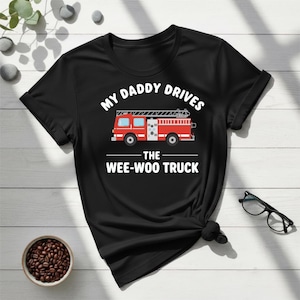 Peut inclure: T-shirt noir avec l'inscription "MY DADDY DRIVES THE WEE-WOO TRUCK" au-dessus d'un dessin de camion de pompiers rouge. Le t-shirt est en matière souple et convient au quotidien.