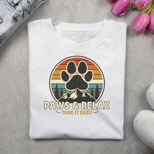 Puede incluir: Camiseta blanca con un diseño circular que presenta una huella de pata negra, montañas y pinos. El diseño incluye el texto "PAWS & RELAX - TAKE IT EASY" en un estilo vintage. La camiseta está doblada sobre una superficie gris.