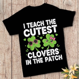 Peut inclure: T-shirt noir avec l'inscription "I TEACH THE CUTEST CLOVERS IN THE PATCH". Le motif présente deux trèfles de dessin animé, l'un portant des lunettes de soleil et l'autre un nœud rose. Des cœurs et des accents verts complètent le thème de la Saint-Patrick.