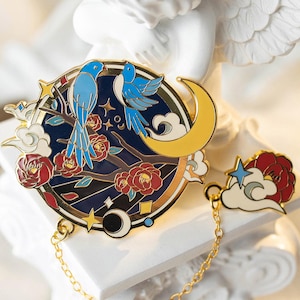 Op de afbeelding: Een ronde emaille pin met twee blauwe vogels op een tak met rode bloemen, een gele maan en een sterrenhemel ontwerp. De pin is verbonden door een gouden ketting met een wolk en bloem charme.