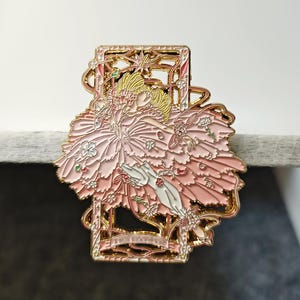 Può includere: Spilla in smalto rosa raffigurante una ballerina in un abito floreale, incorniciata da un bordo decorativo dorato. La spilla presenta un design dettagliato con sfondo rosa chiaro e dettagli bianchi e verdi.
