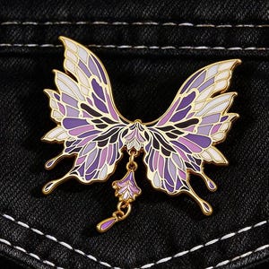 May include: A butterfly-shaped enamel pin with gold-coloured trim. The wings are a gradient of purple and white, with black detailing. A small, dangling charm hangs from the centre. The pin is set against a black denim background.