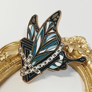 May include: A butterfly-shaped enamel pin with a detailed design. The pin features teal, white, and black wings with gold accents. A silver chain wraps around a black rectangular shape. The pin is set against a gold-colored decorative frame.