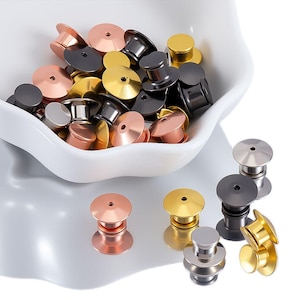 May include: A collection of pin backs in various metallic finishes, including gold, silver, copper, and black. The pin backs are in a white bowl and scattered on a white surface. The pin backs are used for attaching pins and badges.