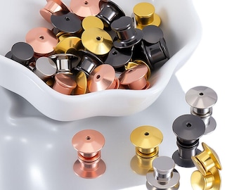Op de afbeelding: Een verzameling pinruggen in verschillende metallic afwerkingen, waaronder goud, zilver, koper en zwart. De pinruggen liggen in een witte kom en zijn verspreid over een wit oppervlak. De pinruggen worden gebruikt voor het bevestigen van pins en badges.