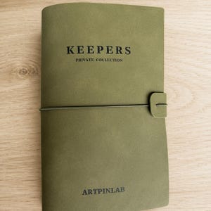 May include: An olive green notebook with a smooth matte finish, featuring a horizontal elastic band closure. The cover is embossed with the text KEEPERS PRIVATE COLLECTION at the top and the brand name ARTPINLAB printed at the bottom.