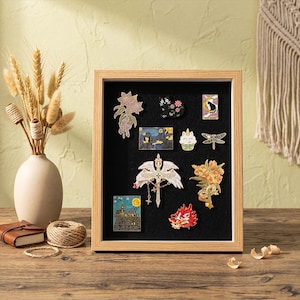 May include: A wooden framed display case with a black felt interior, showcasing various enamel pins. The pins feature designs such as a black cat, a starry night scene, a dragon, and floral motifs. The case is set on a wooden surface.