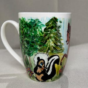 May include: White ceramic mug with a hand-painted forest scene. The design features a skunk, deer, and trees. The mug has a curved handle and a colourful illustration of a woodland setting. The design includes green, blue, brown, black, and white colours.