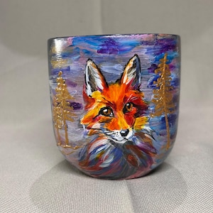 Puede incluir: Taza de cerámica pintada a mano con un retrato de zorro. El zorro tiene pelaje naranja, ojos marrones y hocico blanco, sobre un fondo de cielo colorido y árboles dorados. Una pieza de arte única.