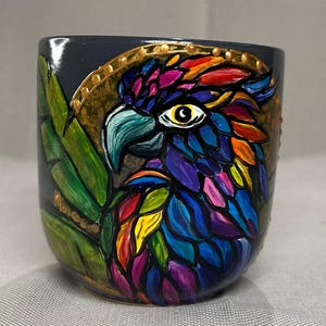 May include: A hand-painted ceramic mug featuring a colorful parrot design. The parrot's feathers are depicted in vibrant hues of red, orange, yellow, green, blue, and purple. The mug has a dark gray background with gold accents and green leaves.