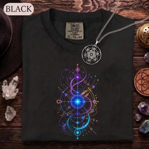 May include: A black t-shirt featuring a vibrant, multicolored geometric graphic print with circular and linear patterns in shades of blue, purple, and orange. The shirt includes a Comfort Colors brand label at the neck, size XL, and a silver pendant.