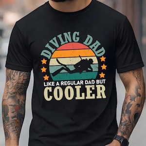 May include: A black t-shirt featuring a graphic of a scuba diver silhouette centered within a sunset-colored circle. The text reads DIVING DAD LIKE A REGULAR DAD BUT COOLER, surrounded by small orange stars in a distressed, vintage-style print.