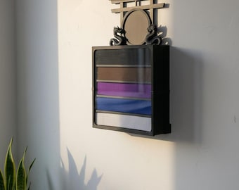 Pode incluir: Uma vitrine de parede preta com frente de vidro e cinco compartimentos horizontais contendo faixas de artes marciais dobradas nas cores preta, marrom, roxa, azul e branca. O topo é adornado com um portal torii e dois dragões.