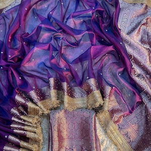 Puede incluir: Una tela transparente con degradado morado y azul que presenta pequeños adornos blancos dispersos. Los bordes están rematados con una ancha y compleja cenefa de color oro metálico con motivos florales y un delicado fleco inferior.
