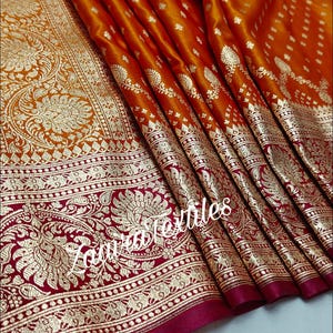 May include: Close-up of a draped orange and red textile with intricate gold floral patterns. The fabric features a detailed border with gold designs on a deep red background. The text "Zauratextiles" is visible.