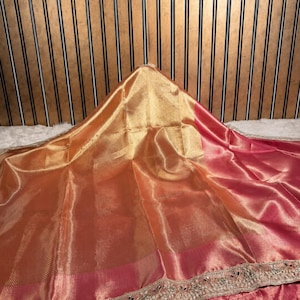 Puede incluir: Un drapeado de tela en color melocotón y dorado brillante que presenta un borde ancho con intrincados bordados plateados, detalles de hilo metálico y pequeños acentos reflectantes a lo largo del borde inferior, con una textura suave y lustrosa.