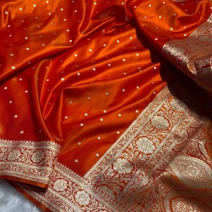 May include: Vibrant orange silk saree with a delicate pattern of small white dots. The saree features intricate gold floral borders and a rich, flowing texture, showcasing traditional craftsmanship.