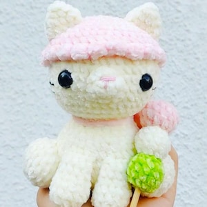 May include: A handmade white crochet cat wearing a pink hat and collar. The cat has black eyes and a pink nose. It holds a green and pink pom-pom skewer. The cat is a cute, whimsical toy.