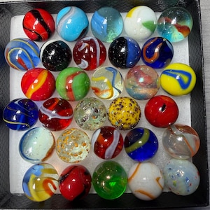 May include: A collection of twenty-five glass marbles arranged in a square grid within a black box. The marbles feature various designs, including solid colors, swirling patterns, speckled textures, and clear glass with internal colored ribbons.