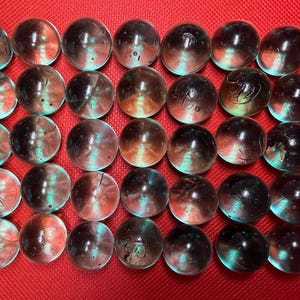 May include: A collection of thirty-two glass marbles, each with a unique blend of colors. The marbles feature a mix of brown, red, and teal hues, creating a captivating visual effect. The marbles are arranged on a red surface.