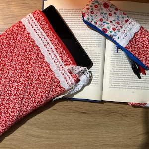 Pode incluir: Capa para tablet e estojo para canetas em tecido floral vermelho com acabamento em renda branca. A capa do tablet é acolchoada e tem uma etiqueta branca que diz "Fil d'Amour". O estojo para canetas tem um zíper azul.