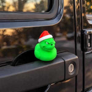 May include: A bright green rubber duck figure with a grumpy facial expression, wearing a small red and white Santa hat. The figure is positioned on the black door handle of a vehicle, showcasing a smooth texture and distinct character features.