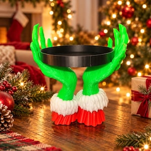 Puede incluir: Un soporte decorativo con dos manos de color verde brillante con dedos largos que se extienden hacia arriba para sostener una bandeja circular negra. Las muñecas tienen puños de piel blanca y mangas rojas plisadas.