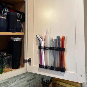 Puede incluir: Un armario de cocina abierto con varios vasos, tazas y un organizador de metal negro que contiene múltiples pajitas reutilizables de diferentes colores. La puerta del armario es de color blanquecino y el interior tiene estantes con artículos para beber.