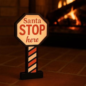 May include: A decorative Christmas sign in the shape of a stop sign. The sign is black with a white and red striped pole. The sign reads "Santa STOP here" in red letters. The background is a cozy fireplace.