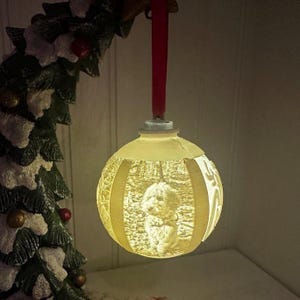May include: A glowing, off-white Christmas ornament with a detailed relief of a dog. The ornament is round with vertical cutouts and hangs from a red ribbon. A Christmas tree is visible in the background.