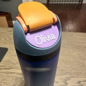 Könnte beinhalten: Eine blaue Trinkflasche mit dunkelblauem Korpus und grauem Deckel. Der Deckel verfügt über einen leuchtend orangefarbenen Klappverschluss und einen strukturierten, hellvioletten runden Einsatz auf der Vorderseite mit dem Namen Olivia in weißer Schrift.
