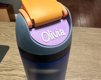 May include: A blue water bottle featuring a dark blue base and a gray lid assembly. The lid includes a bright orange flip-top cap and a textured, light purple circular insert on the front that displays the name Olivia in white raised lettering.