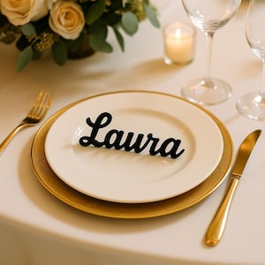 May include: A formal table setting with a white plate and gold charger. A black cursive nameplate that reads "Laura" sits on the plate. A gold fork and knife are on the table, along with wine glasses and a candle.