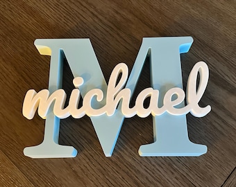 May include: A light blue wooden letter "M" with the name "michael" in white script. The wooden letter has a light blue face with yellow edges. The name "michael" is in a cursive font and is layered on top of the letter.