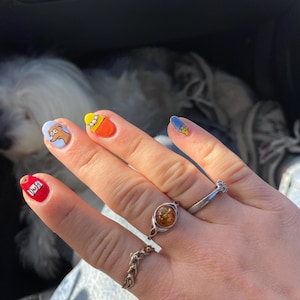 Puede incluir: Una mano con uñas pintadas con diseños de personajes de dibujos animados. Las uñas están pintadas en varios colores, incluyendo rojo, amarillo y azul, con diseños de personajes de dibujos animados. La mano también tiene varios anillos.