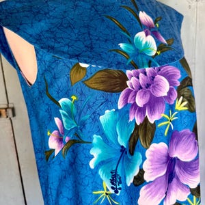 May include: A sleeveless blue dress featuring a dark blue crackle pattern throughout the fabric. The garment is decorated with a vibrant floral print consisting of large purple and light blue hibiscus flowers, green leaves, and small yellow accents.