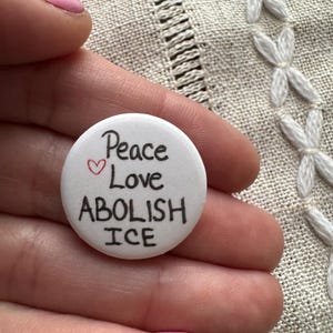 May include: A white button with the words "Peace Love ABOLISH ICE" in black lettering and a small red heart. The button is held in a hand with pink nail polish.